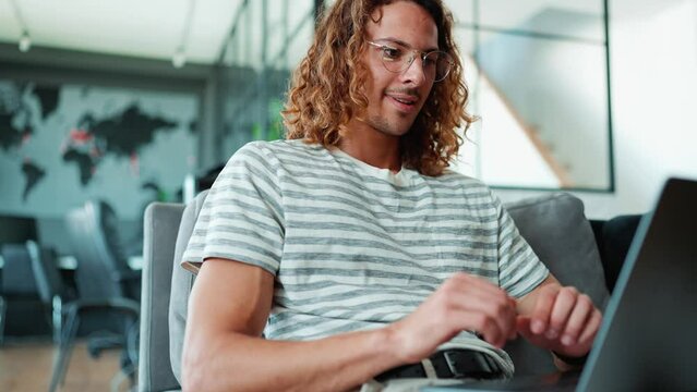 Cheerful curly long haired man in eyeglasses working on laptop in office  