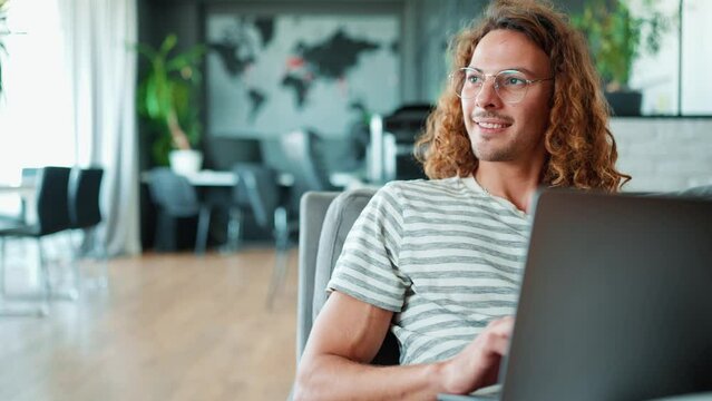 Smiling curly long haired man in eyeglasses working on laptop in office  