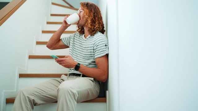 Cheerful curly haired man in eyeglasses texting on mobile and drinking coffee on stairs in office