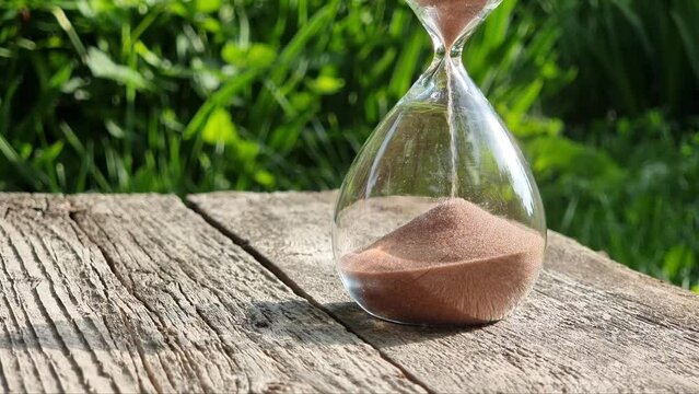 Glass hourglass on a wooden table on a blurred green background. Clear summer day. The concept of time and eternity.