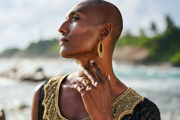 Trans sexual ethnic fashion model in long posh dress and accessories in elegant posture touches neck. Epatage gay black man in luxury jewelry poses, touches neck on scenic ocean beach. Pride, close up