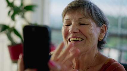 Happy older woman waving hello or goodbye while holding cellphone device at home speaking by video...