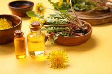 Bottles with cosmetic oil and bowls of dandelion flowers on yellow background