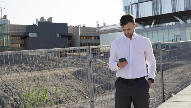 Young businessman using smartphone while walking near construction area. Professional male employer typing text message and talking on mobile phone outside business building construction area.