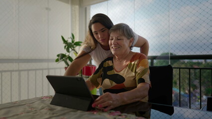 Diverse grand-daughter helping her caucasian grandmother to use tablet device at apartment balcony. Generational family scene teaching older woman to use modern technology