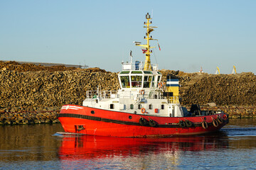 Powerful tugboat with a red hull in harbor channel on wooden logs pile and blue sky background