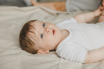 Charming blue-eyed 6-month-old baby lies in bed in a white bodysuit. View from above