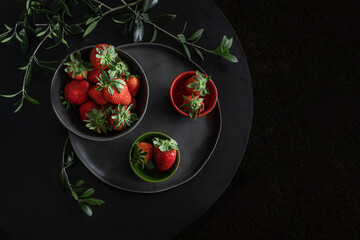 Shot of the fresh strawberries lying in a black handmade cup and plate on a black round table surrounded by green twigs, top view