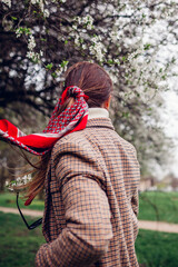 Stylish young woman wears red hair scarf in spring park by blooming trees. Retro female fashion. Accent headscarf.