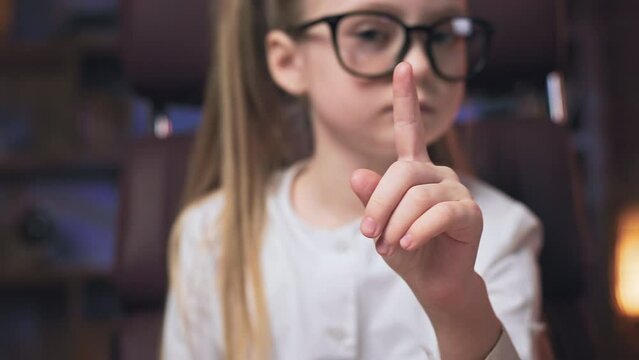 Crop of serious small girl looking at camera and waving index finger to say no. Clever preteen kid in glasses sitting in rolling office chair, shaking head and making denial gesture.