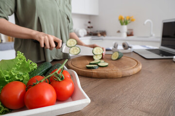 Woman cutting cucumber for salad on wooden table with vegetables