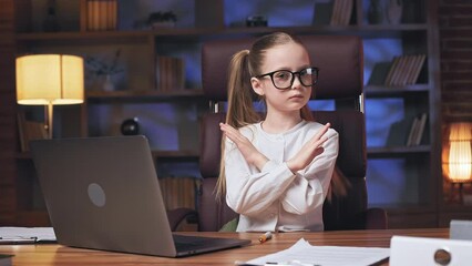 Pretty little girl with serious facial expression showing stop gesture with crossed hands and looking at camera. Confident school child sitting at modern office desk with wireless laptop. - Powered by Adobe