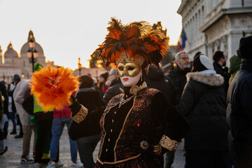 Venice, Italy, 11 February 2023: Colorful carnival masks at famous traditional festival on Saint Mark's Square at sunset, Beautiful Elegant Venetian Costume, black velvet dress with orange feathers
