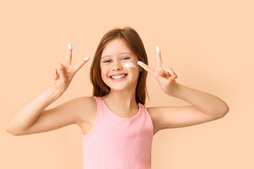 Little girl with sunscreen cream on her face showing victory gesture against beige background