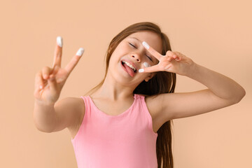 Fototapeta premium Little girl with sunscreen cream on her face showing victory gesture against beige background
