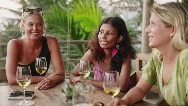 Diverse female friends drink wine in rooftop sea view restaurant at sunset. Multiracial women talk, smile in tropical cafe at dusk. Young multiethnic women dining. Palm trees, ocean in background.