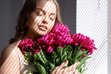 Beautiful young woman with bouquet of peony flowers near window with blinds