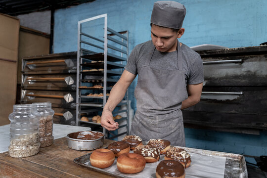 A young Hispanic baker is dropping almond toppings on a chocolate donut