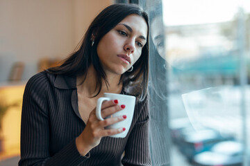 Beautiful thoughtful woman drinking a cup of coffee while looking forward next to the window at home