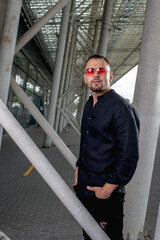 a stylish man in light pants and a black shirt against the background of empty walls holds glasses in his hands and looks away angrily