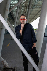 a stylish man in light pants and a black shirt against the background of empty walls holds glasses in his hands and looks away angrily