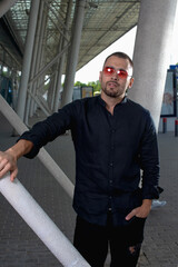 a stylish man in light pants and a black shirt against the background of empty walls holds glasses in his hands and looks away angrily