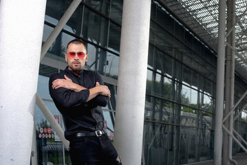 a stylish man in light pants and a black shirt against the background of empty walls holds glasses in his hands and looks away angrily