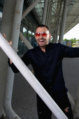 a stylish man in light pants and a black shirt against the background of empty walls holds glasses in his hands and looks away angrily