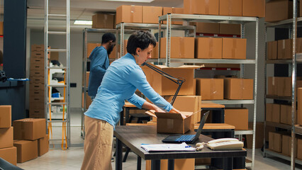 Young startup owner putting merchandise in boxes, working with laptop to do goods inventory before shipping products to clients. Female worker planning delivery management and logistics.