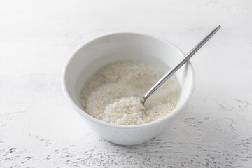 White bowl with long grain rice soaked in water on a light gray background. Cooking delicious steamed rice