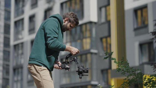 Young professional videographer with camera in his hands shoots video against background of high-rise buildings in residential complex. Backstage. Slow motion