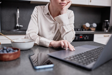 Closeup of young woman using laptop at kitchen table and scrolling work messages, copy space