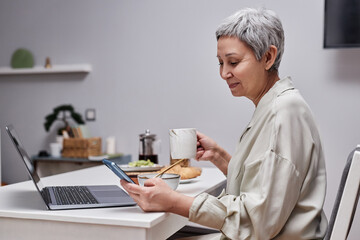 Side view portrait of modern senior woman using smartphone and working while enjoying breakfast in morning
