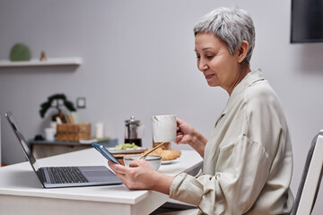 Side view portrait of modern senior woman using smartphone and working while enjoying breakfast in...