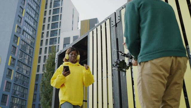 Behind scenes of creating video content, professional cameraman shoots african american girl in yellow hoodie with headphones on territory of residential complex. Backstage. Slow motion