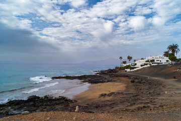 Fototapeta na wymiar Black beach in the bay of the resort town named Puerto del Carmen. Beach lines and the traditional white-washed houses. Lanzarote, Canary Islands, Spain