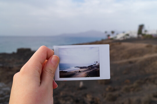 A young woman's hand holds a polaroid photo in front of the view where it was taken