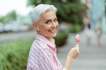 Cheerful stylish gray haired woman looking at camera while eating tasty ice cream in park
