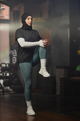 Young active female sports trainer in hijab and activewear doing exercise for leg muscles in leisure center while keeping left one bent in knee