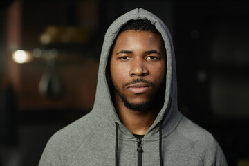 Serious African American male fitness instructor in grey activewear with hood standing in front of camera in gym and looking at you