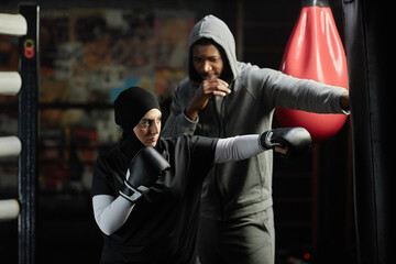 Young Muslim woman in activewear, hijab and boxing gloves hitting black leather punching bag while...