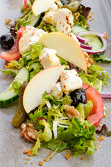 Close-up of a fresh salad with grilled goat cheese, apple, celery, walnuts, tomato, red onion, lettuce, cucumber, pine nuts, walnuts, olives and pickle topped with a honey dressing on a gray plate.