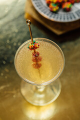 golden cocktail pick in glass with colorful clusters of candy, reflective metallic gold background