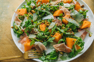 top down view of arugula salad with cheese, cantaloupe, prosciutto, pistachios on white plate....