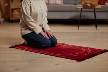 Cropped shot of young Islamic woman in casualwear kneeling on small red rug on in living room while...