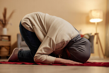 Side view of young Muslim woman in traditional hijab bending forwards while kneeling on small red...