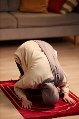 Young Muslim woman in beige pullover and grey hijab praying on small red rug while kneeling on the...