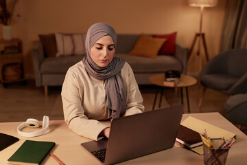 Young serious Muslim businesswoman sitting by workplace in living room and looking at laptop screen while scrolling through online data