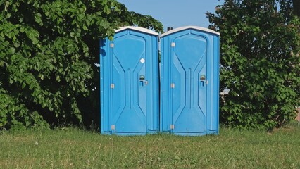 Pair of Blue Plastic Mobile Public Toilet Outhouse Standing in the Middle of High Grass Meadow...