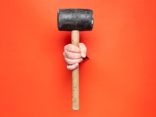 A hand is holding a big rubber mallet through torn orange paper. No face, copy space, concept.
